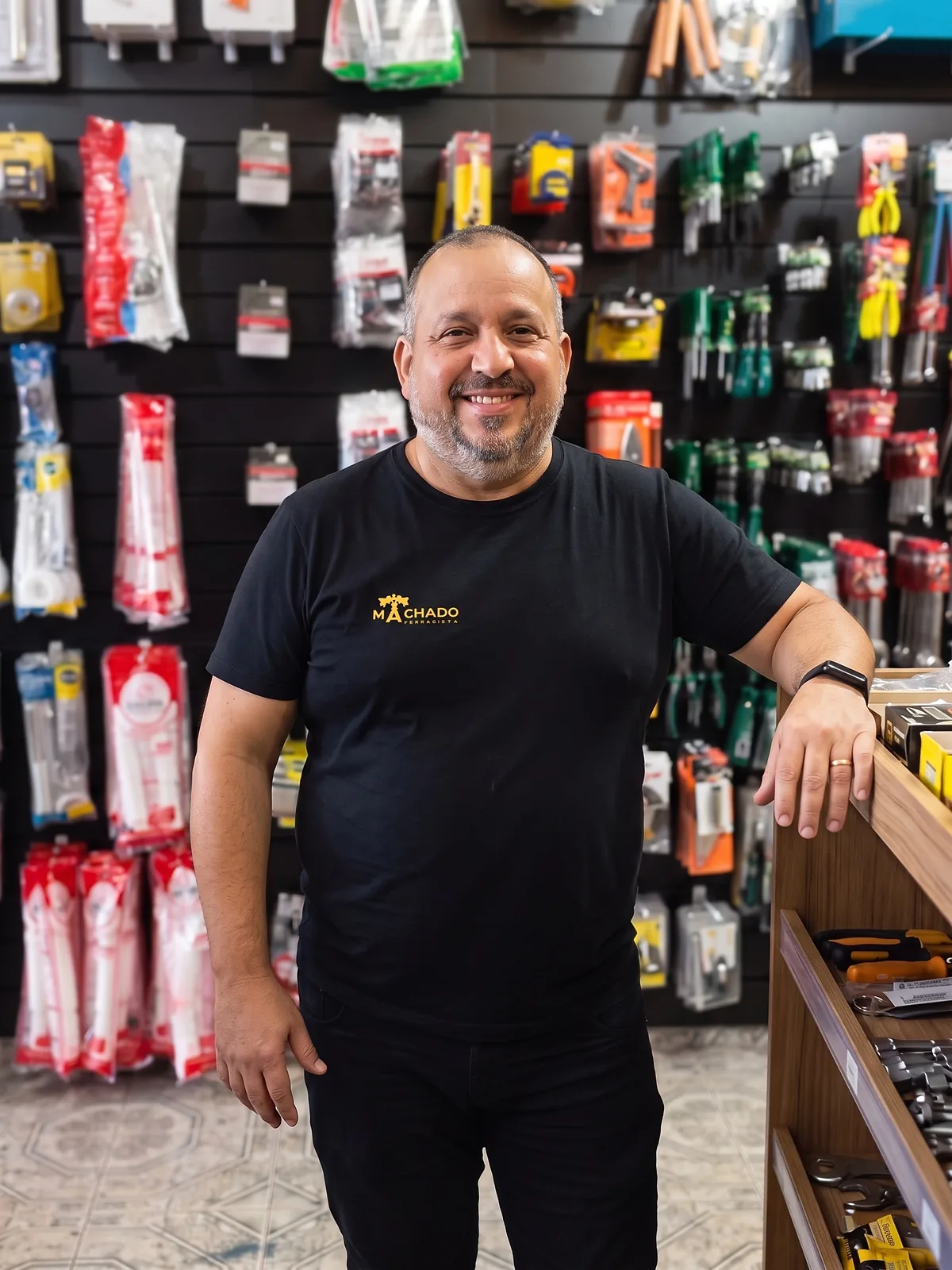 Alexandre Machado, dono da Machado Ferragista, sorrindo dentro da loja diante da parede de ferramentas
